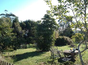 Un jardín soleado con césped verde, varios árboles y arbustos. Sillas de metal y una mesa de madera están dispuestas para relajarse al aire libre. Ambiente tranquilo, natural y acogedor.