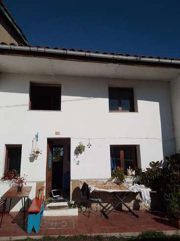 Una casa blanca de dos plantas con tejado de tejas, ventanas abiertas y un patio. Mesas, plantas y un banco decoran el espacio exterior bajo un cielo azul despejado.