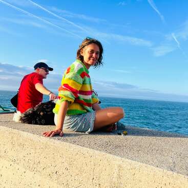 Uma mulher sorridente com um suéter colorido senta-se em um muro de concreto à beira-mar. Um homem e um cachorro preto e branco relaxam nas proximidades, sob um céu azul.