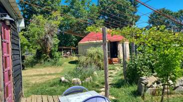In einem sonnigen Garten steht eine Jurte mit rotem Dach, umgeben von üppigem Grün, Sitzgelegenheiten im Freien, einem Holzhäuschen und Aufhängungen für Hängepflanzen.
