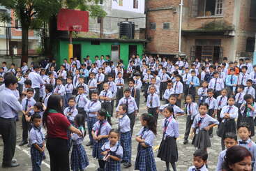 A large group of school children in uniform stand in a courtyard, supervised by teachers, possibly during a morning assembly at a school outdoors.