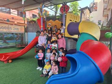 A group of young children and two adults pose together in front of colorful slides at a playground, holding balls, smiling, and enjoying outdoor playtime.