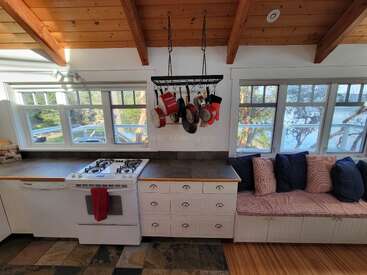 Cette cuisine confortable comprend une cuisinière, des casseroles suspendues, une banquette rembourrée près de la fenêtre, des poutres apparentes, de grandes fenêtres offrant une vue panoramique, des plans de travail carrelés et une lumière naturelle et chaleureuse.