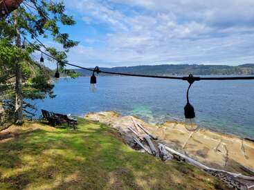 Des guirlandes lumineuses sont suspendues au-dessus d'une falaise verdoyante bordée d'arbres qui surplombe un lac bleu paisible. Des chaises en bois et une table offrent un refuge paisible et pittoresque au bord du lac.