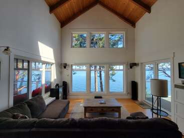 Un salon lumineux et spacieux avec un plafond voûté en bois, de grandes fenêtres donnant sur un lac paisible et des arbres, un mobilier moderne et des éléments de décoration naturels et chaleureux.