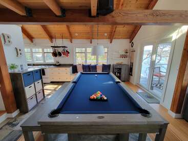 Cette image montre un espace de vie confortable et ensoleillé avec une table de billard bleue au centre, des poutres en bois rustiques, une cuisine et des portes coulissantes donnant sur l'extérieur.