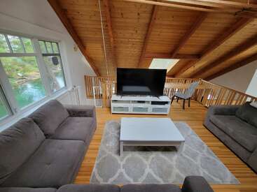 Ce salon loft confortable dispose d'un plafond en bois incliné, de grandes fenêtres avec vue sur l'eau, de canapés gris, d'une télévision, d'une table blanche et d'un tapis à motifs.