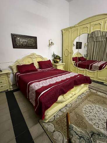 Dieses Schlafzimmer ist mit kunstvoll verzierten gelben Möbeln, einem großen Bett mit roter Bettdecke, einem Spiegelschrank, Nachttischen, einem Blumengesteck, einem beigen Teppich und islamischer Wandkunst ausgestattet.