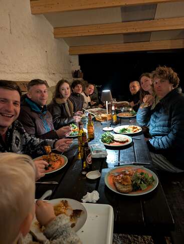 Un groupe de personnes est assis autour d'une table en bois et partage un dîner. Les assiettes, les boissons et une lampe créent une ambiance chaleureuse, conviviale et douillette.