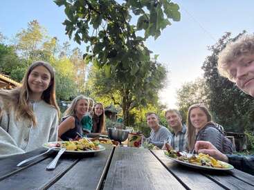 Un grupo de siete personas disfruta de una comida al aire libre en una mesa de madera, rodeadas de árboles y naturaleza, sonriendo con calidez bajo la luz del sol del atardecer.