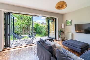 This bright living room features modern furniture, a large sofa, wall-mounted TV, and opens to a lush garden with patio chairs through wide folding glass doors.