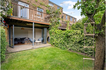 This image shows a cozy backyard garden with green grass, a tree with a rope ladder, a leafy fence, open patio doors, and an upstairs balcony.
