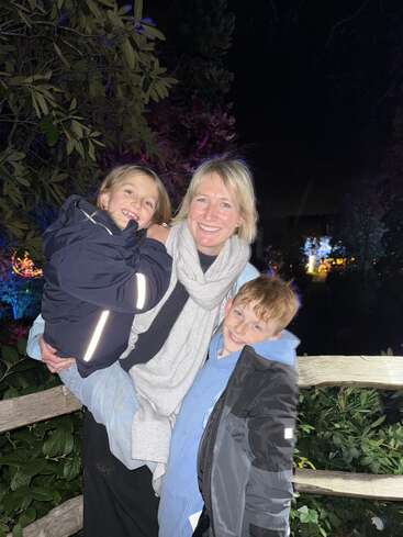 Eine Frau und zwei Kinder posieren nachts im Freien und lächeln dabei. Sie stehen vor einem Holzzaun und belaubten Büschen, die von Lichtern beleuchtet werden.