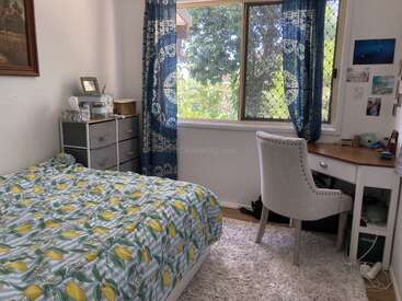 Cette chambre confortable comprend un lit avec une literie à motifs de citron, un tapis blanc doux, une commode, un bureau d'angle, une chaise rembourrée, des rideaux bleus et une grande fenêtre avec vue sur la verdure.
