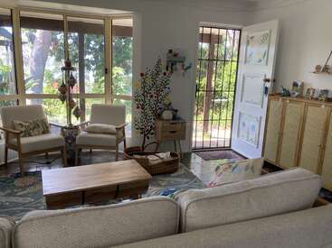 Un salon confortable et ensoleillé avec de grandes fenêtres, deux fauteuils, une table basse en bois, un tapis coloré, un tableau mural représentant un arbre, des coussins et une porte ouverte décorée.