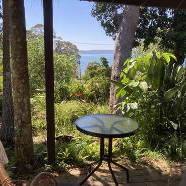 Une petite table en verre est installée sur un patio ombragé entouré d'une végétation luxuriante, avec une vue imprenable sur l'eau bleue et les collines boisées lointaines.