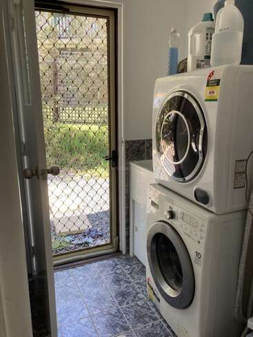 Il s'agit d'une petite buanderie où sont empilés un lave-linge et un sèche-linge. Il y a du détergent sur le dessus, le sol est carrelé et une porte moustiquaire ouverte mène à l'extérieur.