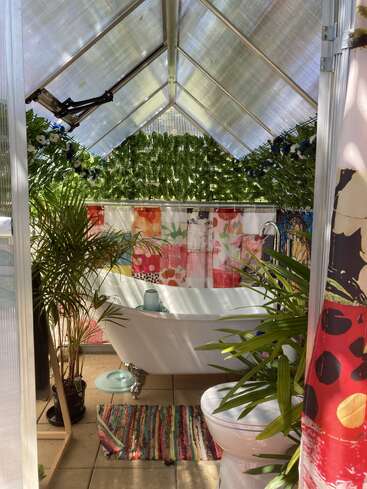 Cette image montre une salle de bains lumineuse, de type serre, avec une baignoire sur pattes blanche, un décor coloré, des plantes vertes luxuriantes, un sol carrelé et une atmosphère vibrante et joyeuse.