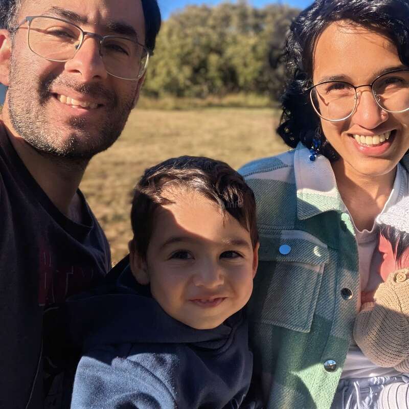 Une famille de quatre personnes - le père, la mère, le jeune garçon et le bébé - sourit chaleureusement à la lumière du soleil, profitant d'une journée en plein air avec des arbres en arrière-plan.