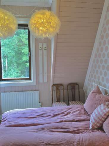 Gemütliches Schlafzimmer mit rosafarbener Bettwäsche, zwei flauschigen Pendelleuchten, drei Holzstühlen, einer gepunkteten Akzentwand, einem Fenster mit grünen Bäumen und einer hellen Holzverkleidung.