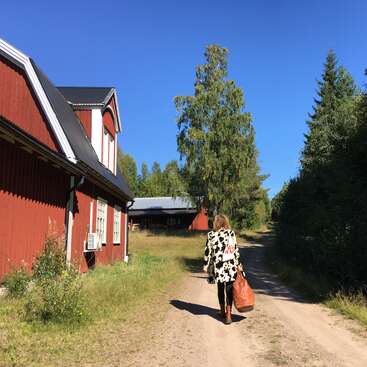 Une femme marche sur un chemin de terre à côté de bâtiments en bois rouge, en portant un sac brun. Les grands arbres et le ciel d'un bleu éclatant créent une atmosphère rurale paisible.