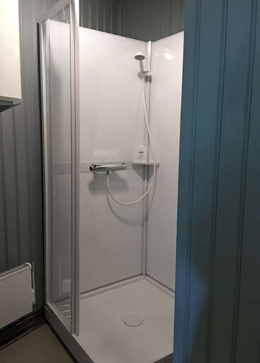 Dieses Bild zeigt eine einfache, moderne Duschkabine mit einer Glastür, weißen Wänden, einer Handbrause und einem kleinen Regal mit einer Seifenflasche.