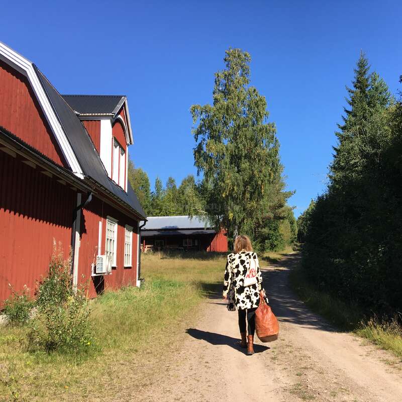 Eine Frau geht mit einer braunen Tasche auf einem Feldweg neben roten Holzhäusern entlang. Hohe Bäume und ein strahlend blauer Himmel schaffen eine friedliche, ländliche Atmosphäre.