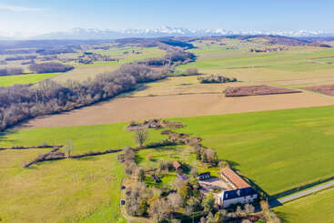 This image shows a beautiful rural landscape with green fields, scattered trees, a farmhouse, and distant snowy mountains under a clear blue sky on a sunny day.