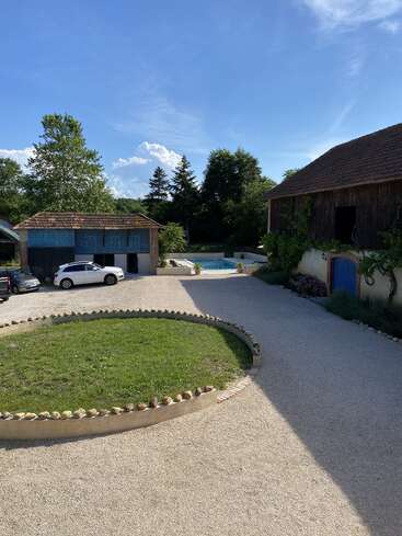 A sunny courtyard with two rustic buildings, a circular grassy area, gravel driveway, two parked cars, surrounded by trees, and a pool in the background.