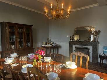 A warmly lit dining room features a wooden table set with bowls, glasses, pink flowers, a chandelier above, ornate mirror, cabinet, and fireplace in the background.