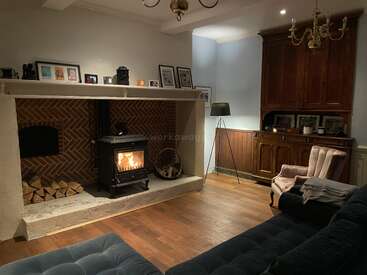 A cozy living room with a burning wood stove, brick fireplace, logs, framed photos, velvet armchair, dark sofa, wooden floors, lamp, and cabinet. Warm ambiance.