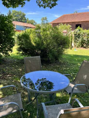 A glass outdoor table with four chairs is set on a grassy lawn, surrounded by bushes and trees, with rustic buildings and clear blue sky in background.