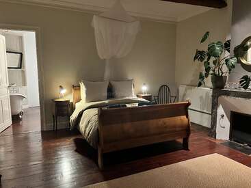 A cozy bedroom with a wooden sleigh bed, bedside lamps, mosquito net, indoor plant, fireplace, armchair, wooden floor, and an adjoining bathroom with a tub.