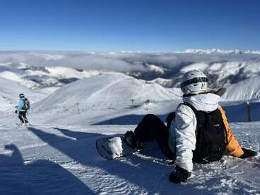 A snowboarder sits on a snowy mountain slope, gazing at the scenic view. Another person skis downhill. The sky is clear, mountains stretch endlessly.