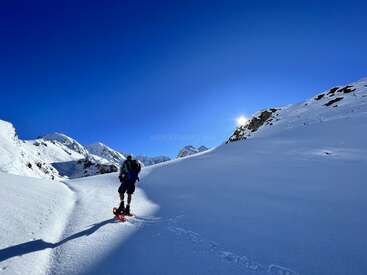 A person in snowshoes hikes along a snowy mountain path under a deep blue sky. Bright sunlight peeks over the ridge, casting long shadows.
