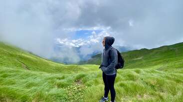 A person stands on lush green hills, wearing a hoodie and backpack, gazing at the cloudy sky. Misty mountains and vibrant grass create a serene atmosphere.