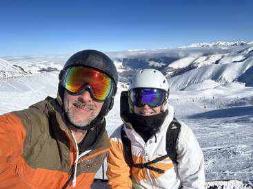 Two people wearing ski gear and helmets smile for a selfie on a snowy mountain. The bright sun reflects off their goggles, surrounded by beautiful winter scenery.