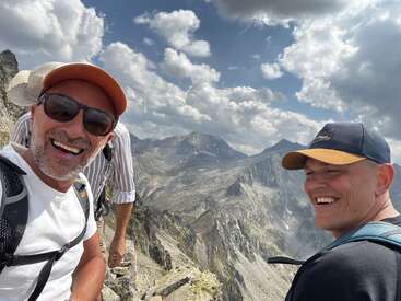 Three smiling men are hiking on a rocky mountain peak under a dramatic cloudy sky, enjoying the stunning scenic landscape and capturing their adventure with a selfie.