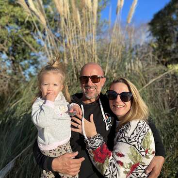 A happy family of three poses outdoors in front of tall grass, all smiling and wearing sunglasses. The sky is clear and blue.