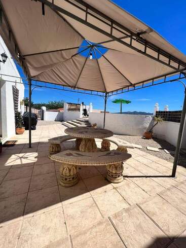 Unter einem beigen Vordach steht ein Steintisch mit verzierten Bänken auf einer gefliesten Terrasse. Der Bereich ist sonnig, mit strahlend blauem Himmel und Pflanzen im Freien.