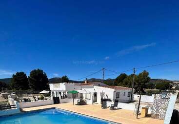 In einem hellen, sonnigen Innenhof gibt es einen Swimmingpool, Gebäude mit weißen Wänden, Liegestühle und einen grünen Sonnenschirm, umgeben von Hügeln, Bäumen und einem strahlend blauen Himmel.