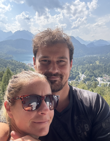 Ein Paar macht ein Selfie im Freien, umgeben von schönen Bergen, Wäldern und einem See. Die Frau trägt eine Sonnenbrille, die den Blick auf die Landschaft reflektiert.