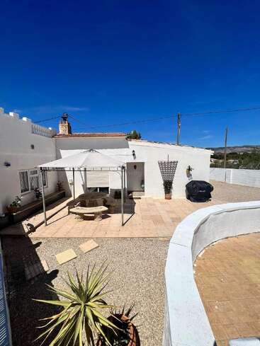 Um pátio ensolarado apresenta um pátio de azulejos, edifícios brancos, uma área de estar à sombra com bancos, vasos de plantas, cascalho, uma churrasqueira e um céu azul brilhante.