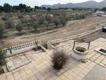 L'image montre un patio carrelé avec un buisson sec et sans feuilles et un puits en pierre. Au-delà, il y a un champ rural avec des arbres épars et des montagnes lointaines.