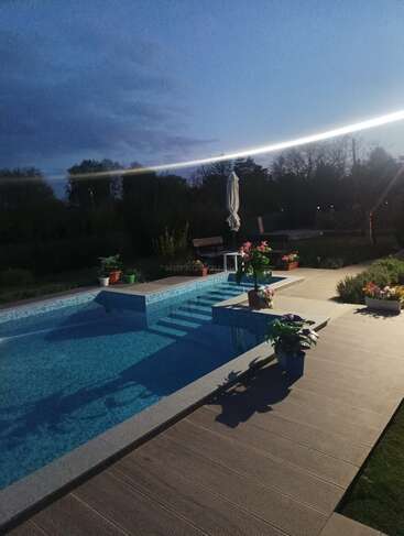 Ein ruhiger Abend am Pool, umgeben von Topfpflanzen und Blumen, mit einem geschlossenen Sonnenschirm, einer Holzterrasse und Bäumen, die sich vor dem dunkler werdenden Himmel abzeichnen.