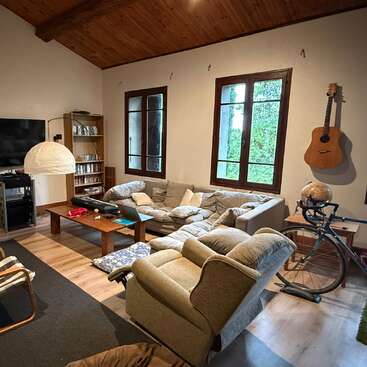 Cozy living room with beige sofas, recliner, wooden ceiling, large windows, guitar on wall, coffee table, bicycle, globe, bookshelf, TV, and indoor greenery views.