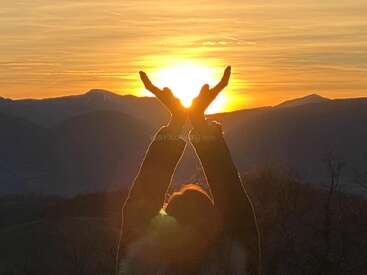 A person stands against a mountain backdrop at sunset, holding their hands up as if cradling the glowing sun, creating a tranquil, uplifting silhouette scene.