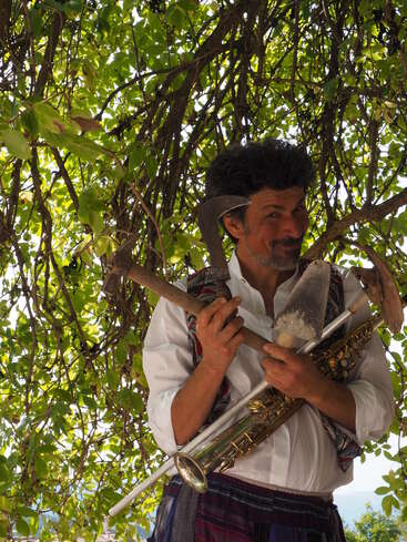 L'image représente un homme debout devant un arbre, tenant une pelle et un saxophone, vêtu d'une chemise blanche et d'un gilet coloré, sur fond de feuilles vertes luxuriantes.