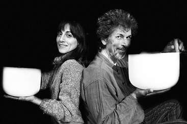A smiling woman and man sit back-to-back, each holding a large white bowl. The image is black and white, emphasizing their joyful expressions and connection.