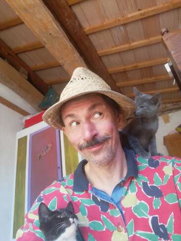 A cheerful man wearing a straw hat and colorful shirt smiles playfully. He holds a black-and-white cat while a gray cat sits on his shoulder indoors.